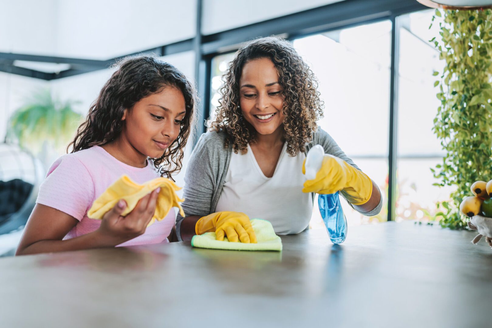 Two women cleaning a table together, smiling and wearing yellow gloves.